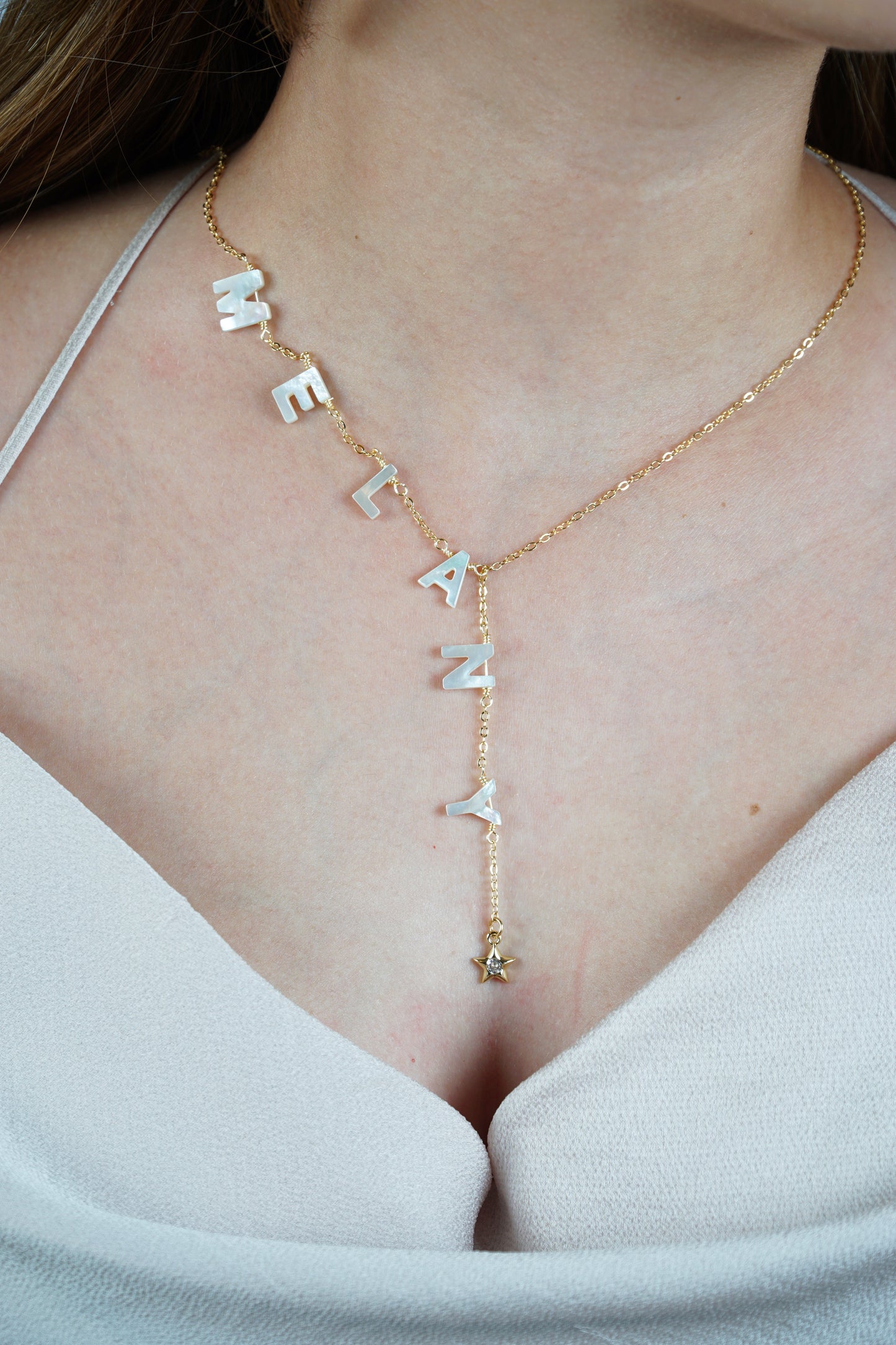 A woman wearing a white v-neck t-shirt, displaying a personalized necklace with mother of pearl letters spelling out the name 'Melany.