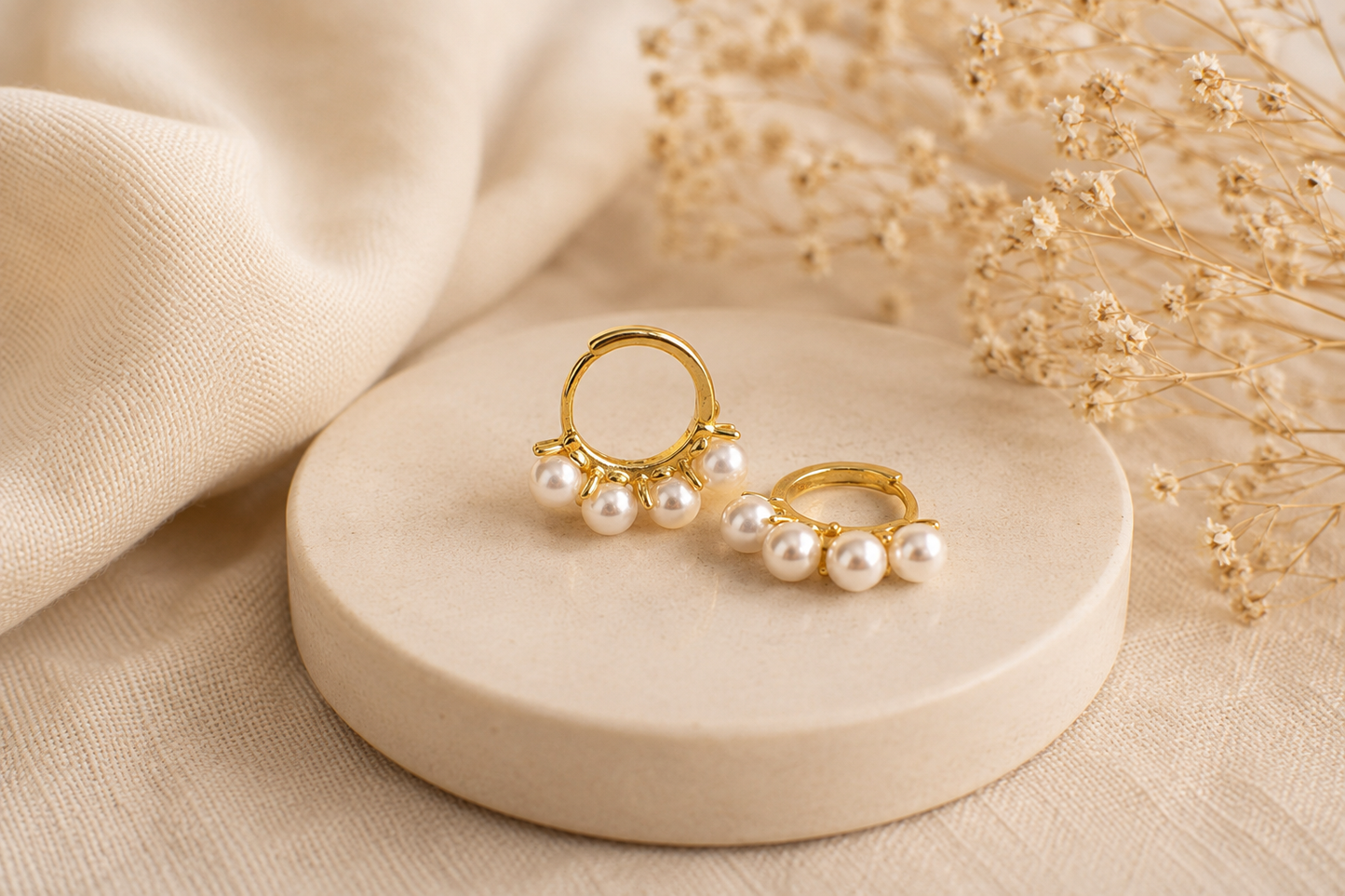 Gold and pearl earrings on a beige stone surface with dried flowers in the background