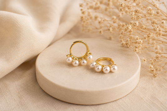 Gold and pearl earrings on a beige stone surface with dried flowers in the background